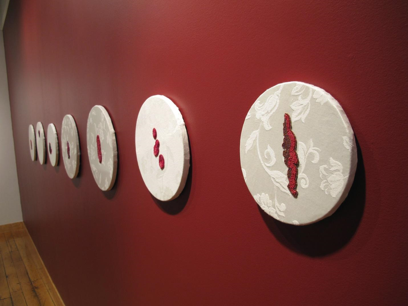 This photograph features “Marked II”, a series of seven beading works,
        they are twelve inch diameter embroideries by Vanessa Dion Fletcher hung
        on a dark red wall in the Tangled Art Gallery as part of her exhibition, <em>Own Your Cervix</em>.
        The circular embroideries are arranged horizontally across the wall. The
        embroidery closest to the camera is more in focus displays a circular frame
        stretched with white damask fabric with a beaded shape similar to a blood
        stain in the centre. Each embroidery work in the series shows different
        shapes of the artists’ period stains embroidered with red beads.