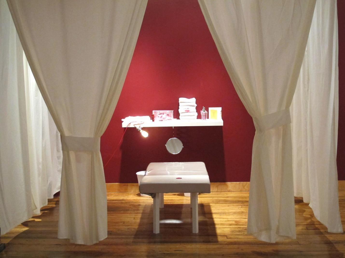 This is a photograph of “Cervical Self Exam Room” an installation by Vanessa
        Dion Fletcher as part of her exhibition Own Your Cervix at Tangled
        Art Gallery. In the centre of the photo, a grey leather examination table
        is arranged in front of a dark red wall. Behind the table on the wall is
        a white floating shelf with white towels folded and stacked in a pile,
        an informational pamphlet, a light bulb, and a circular mirror attached
        to the edge. On the left and right are white curtains that extend from
        the wall and can be drawn to create a private space in which to perform
        self-examinations, in whatever form that might take.