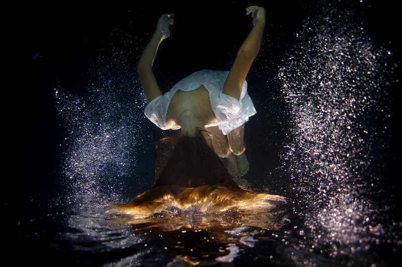 Figure 3. “Underwater_dark01” by Elena Kalis (n.d.). Reprinted
            with permission. A woman wearing a white cotton dress is submerged under
            water. Her body is flipped upside down. In contrast to the darkness of
            the water, tiny white air bubbles surround the woman’s body. Her face cannot
            be seen, only the back of her head and long amber hair.